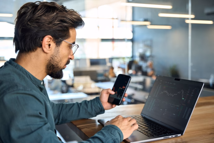 Man with glasses using a laptop and checking stock charts on a smartphone in a modern office.